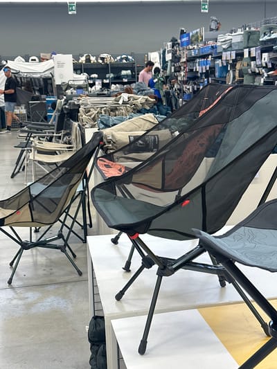 The image shows a retail store selling camping and outdoor gear. In the foreground, several lightweight camping chairs are displayed on white shelving units. These chairs have dark metal frames and fabric seats, some of which are mesh and some solid. One chair in the foreground features a grey fabric seat with a subtle topographic map-like pattern. In the middle ground, more camping chairs are visible, arranged in rows on the floor. Behind these, aisles of merchandise are visible, including tents, sleeping bags, and backpacks, stacked on shelves. Two men are present in the background. One man, wearing a black t-shirt and denim shorts, is walking towards the left of the frame. Another man, wearing a pink t-shirt, is standing further back, looking at merchandise. The environment appears to be a large, well-lit store with concrete flooring and industrial shelving. The overall impression is a retail setting for outdoor equipment. This scene is consistent with a store like Decathlon, which has a location in the San Giovanni Teatino area of Italy.