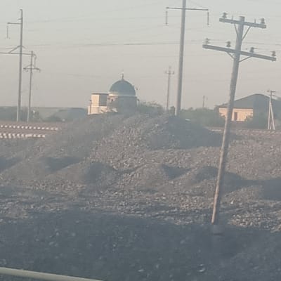 The image depicts a somewhat hazy outdoor scene. The prominent subjects are utility poles with wires crisscrossing the frame, suggesting an infrastructure-heavy environment. In the middle ground, a building with a dome, possibly a mosque or a similar structure, is visible. To the right, a large pile of dark material, likely coal or industrial waste, dominates the landscape. The overall impression is one of a somewhat industrial or semi-rural setting. There are no people visible in the image. The lighting suggests it is daytime, and the haze implies either a dusty atmosphere or overcast weather. Specific location cues are limited, but the provided context places it in Arys, Kazakhstan. There is no visible text in the image.