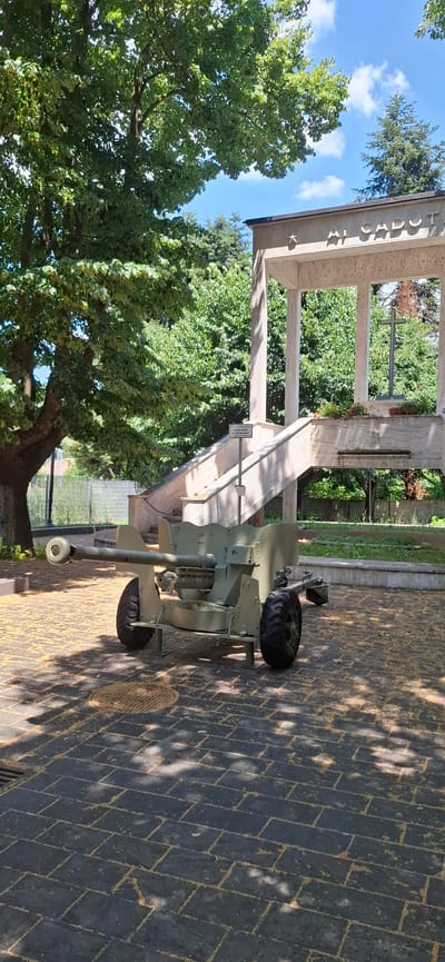 A green artillery piece is displayed on a paved area in front of a war memorial. The memorial features a large concrete structure with columns and a platform, inscribed with "AI CADUTI" which translates to "To the Fallen." A stone staircase leads up to the memorial. The area is surrounded by trees and a clear blue sky with scattered clouds. The ground is paved with dark rectangular stones, and there are some scattered yellow petals or leaves on the ground. A metal fence is visible in the background to the left. A small sign in front of the staircase reads "vietato arrampicarsi sul monumento," meaning "forbidden to climb on the monument." The artillery piece has two large wheels and a barrel pointed towards the left.