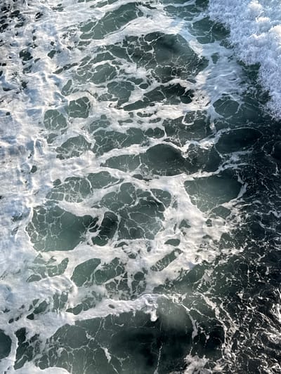 The image depicts a detailed, overhead view of turbulent seawater in Palaiopoli, Greece. The scene is dominated by a dynamic interplay of dark teal-green water and extensive bright white sea foam. The foam forms intricate, irregular patterns across the water's surface, creating a web-like texture, particularly concentrated on the left and central portions of the frame. Towards the right side, the water appears darker, with a pronounced, frothy crest of a wave visible along the upper right edge, indicating active wave action. Light reflects off the foamy surface, creating highlights and contributing to the textured appearance. No individuals, man-made objects, or identifiable land features are present, focusing entirely on the agitated maritime environment.