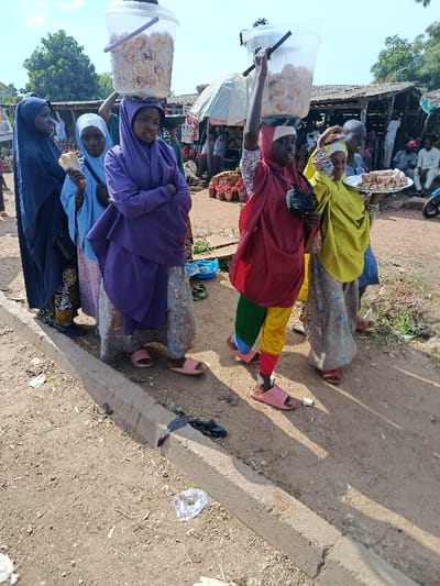 The image depicts an outdoor market scene in Jere North, Nigeria, under clear daylight. Five females are prominently featured in the foreground, standing or walking on a dirt path adjacent to a concrete curb. From left to right: a woman in a dark blue hijab and patterned attire holds a light-colored bread item; a girl in a light blue hijab and patterned dress carries a black shoulder bag and looks towards the camera; a woman in a purple hijab and patterned trousers balances a large, clear plastic bucket filled with circular baked goods on her head, her arms crossed; another woman in a red top, multi-colored (red, green, yellow, blue) trousers, and a black shoulder bag carries a similar clear plastic bucket of baked goods on her head, steadying it with her raised left hand; and a woman in a yellow hijab and patterned dress carries a white circular tray holding light-colored rectangular sweets at chest height. All individuals are wearing sandals or flip-flops. The foreground ground consists of packed dirt with scattered debris, including plastic and paper, near the concrete curb. In the midground and background, multiple rustic market stalls with wooden frames and simple roofs are visible, displaying various goods, including red produce. Several other individuals are present in the blurred background, some seated. A blue scooter is partially visible in the far background. Green foliage from trees is present in the upper left and right background.