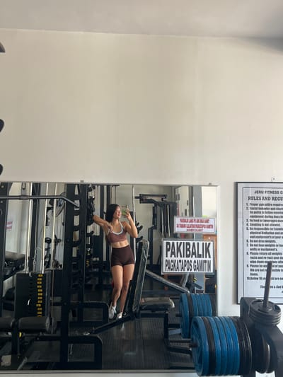 The image depicts a young woman in athletic wear taking a mirror selfie in a gym. She is wearing a brown sports bra and matching shorts, with white athletic shoes. She is leaning against a weight rack, with a smartphone in her right hand. The gym appears to be well-equipped with various weight machines and free weights, including stacks of weight plates in blue and black. A large mirror reflects the gym equipment and the woman. There are signs on the mirror and on the wall. One sign on the mirror reads "PAKI BALIK LAMANG PO ANG MGA GAMIT SA TAMANG LALAGYAN PAGKATAPOS GAMITIN MARAMING SALAMAT!", which translates to "Please return the items to their proper place after use. Thank you very much!". Another sign on the wall is a list of gym rules, with the heading "JERU FITNESS CENTER RULES AND REGULATIONS". The overall environment suggests a functional gym space. The context provided places this scene in the city of Cuyo, Philippines.