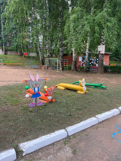 In the city of Sovetsko-Nikolskoye, Russia, an outdoor playground scene is depicted. The foreground features a whimsical wooden sculpture of a rabbit holding colorful balls, standing in front of a carved orange fox sculpture. Further back, a yellow log animal and a green crocodile-like sculpture are visible on the grass. Behind these, a dirt area is marked by a low white concrete curb. The background is dominated by birch trees with lush green foliage, casting dappled shade. A brick building with windows and a basketball hoop is partially visible amongst the trees, along with playground equipment like a soccer goal and a climbing frame. The overall impression is one of a well-used, if slightly worn, children's play area.