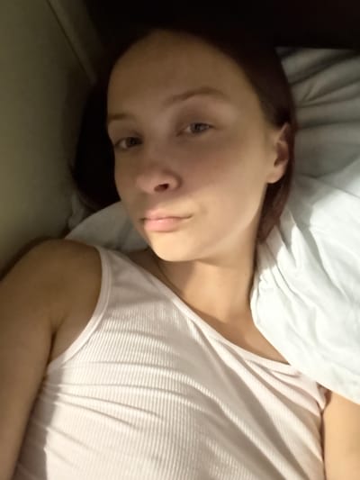 The image shows a close-up, rotated shot of a young person lying on a bed. The subject has brown hair and is looking towards the camera with a neutral expression. They are wearing a light-colored, ribbed t-shirt. A white pillow is visible behind their head and shoulder. The lighting suggests it might be daytime, possibly morning or late afternoon, with soft light casting shadows on the t-shirt. The setting appears to be a bedroom. There is no visible text in the image.