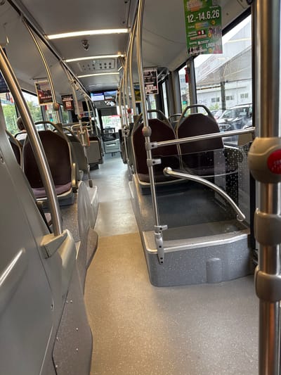 The image shows the interior of a public bus in Liestal, Switzerland. The bus is mostly empty, with rows of brown padded seats along the sides of the aisle. Silver metal handrails run along the seats and overhead. Advertisements are visible on the windows and seat backs. Outside the windows, buildings and a street scene can be seen.