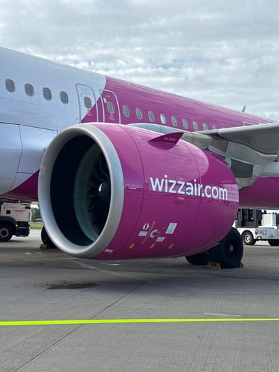An airplane engine, painted magenta with "wizzair.com" written on it in white letters, is visible in the foreground. The engine is large and cylindrical with visible fan blades inside. The fuselage of the airplane, white with a magenta stripe, extends to the left. Several windows are visible on the fuselage. A yellow caution tape runs across the concrete tarmac in front of the plane. In the background, a white utility vehicle with "HL2" on its side is parked to the right of the engine. To the left of the engine, a service truck is parked. The sky is cloudy. The scene appears to be at an airport in Künersberg, Germany.