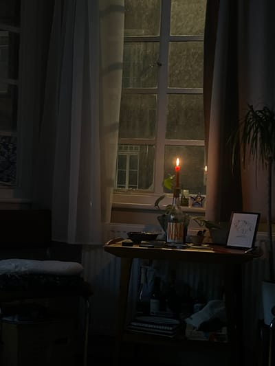 A dimly lit room features a window covered by sheer white curtains. A lit candle sits on a shelf adjacent to the window, along with a framed picture and various objects. The shelf's surface is cluttered. A houseplant is partially visible above the window, and a portion of a chair is seen in the foreground, adding to the cluttered, cozy feel of the interior.