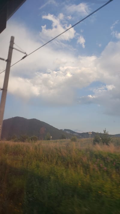 An image captures a view from a moving vehicle, likely a train, looking out towards a landscape. In the foreground, a field of tall grass, blurred due to motion, displays hues of green, yellow, and some scattered purple flowers. A portion of a utility pole, a standard electrical pylon, is visible on the left side of the frame, running vertically. Overhead, two dark wires stretch diagonally across the sky. The sky itself is a mix of bright blue and large, fluffy white clouds, with a hint of a rainbow or light refraction appearing in the upper right quadrant. In the mid-ground, a dark, forested mountain range dominates the horizon. Further back, a less distinct range of hills is visible, with some structures, possibly buildings or industrial elements, emitting faint plumes of smoke. The image appears to be taken in the vicinity of Lučivná, Slovakia, given the characteristic mountainous terrain and vegetation.