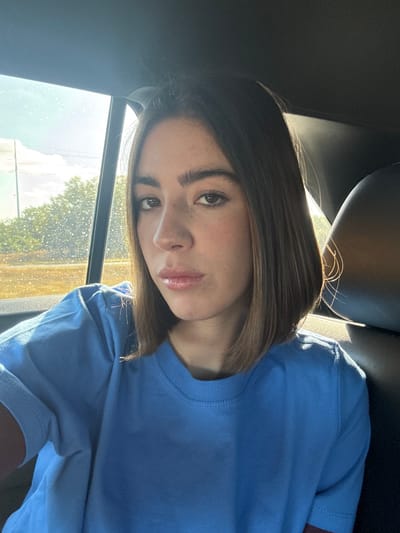 The image shows a single female individual seated in the rear passenger compartment of a vehicle, in the city of Pleasanton, United States. The subject, appearing as a young adult or adult, has medium-length brown hair, dark eyes, and is looking directly towards the camera. She is wearing a light blue, short-sleeved t-shirt. Sunlight streams into the vehicle from the left through a side window. The vehicle's interior, including a visible headrest to the subject's right, is dark. Through the window, an outdoor environment is visible, comprising green foliage, a utility pole with lines, and a bright sky, suggesting daytime. The window glass appears to have dust or water marks. No specific actions or interactions beyond posing for the photo are discernible.