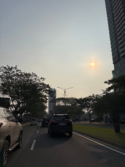 The image captures a street scene in Bencongan Indah, Indonesia, likely during a hazy sunset or sunrise due to the diffused sunlight. A multi-lane road dominates the foreground with several vehicles in transit. On the left, a beige SUV is partially visible. In the center lane, a black SUV is prominent, followed by other cars. Trees line the right side of the road, interspersed with a grassy median and a tall, modern building. On the left, another tall building is visible in the distance. The sky is overcast with the sun appearing as a bright orb, suggesting the atmospheric haze. Streetlights are also present along the road.