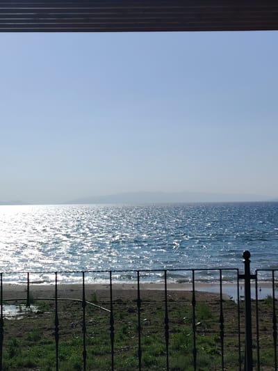 The image presents a bright, sunlit view overlooking a large body of water, identified as Lake Iznik in Turkey. In the immediate foreground, a dark, ornate wrought iron fence with decorative scrolls and vertical bars extends across the lower portion of the frame. Behind the fence, a patch of uneven ground features sparse green grass and some bare earth, leading to a narrow, light-colored sandy beach.

The central and most prominent feature is the expansive blue water, stretching towards the horizon. Its surface is intensely illuminated by direct sunlight, creating numerous dazzling, sparkling reflections that shimmer across the water, indicating strong sun exposure and possibly a slight breeze. In the distant background, faint, hazy outlines of mountains or hills are visible, blending softly with the horizon.

Above the distant landmass, the sky is a clear, uniform light blue, suggesting fair weather conditions. A dark, horizontal structure, resembling an overhang or roof eave, spans the very top of the image. The scene is devoid of people or discernible activity. The strong sunlight and high sky indicate it is likely midday or early afternoon. No visible text is present in the media. The overall impression is a serene and scenic natural landscape.
