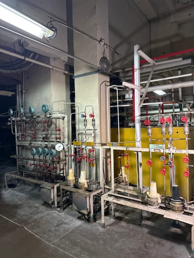 This image displays an interior view of an abandoned industrial facility, located in Чорнобиль, Ukraine, likely within the Chernobyl Exclusion Zone. The scene features a complex network of industrial process equipment, primarily composed of metal piping, numerous red-handled gate valves, and multiple circular pressure or flow gauges. These components are organized across two freestanding, multi-tiered metal racks. The left rack exhibits several gauges at varying heights, including one with a visible dial indicating a near-zero reading. The right rack features an array of vertical pipes equipped with individual red-handled valves and clear, tubular flow indicators. Behind the right-hand equipment, large yellow-painted panels or sections of machinery are visible. The structure of the room includes two prominent, light-colored concrete structural columns. Illumination is provided by a long, overhead fluorescent light fixture and several older, round industrial lamps mounted on the columns and ceiling. The concrete floor beneath the equipment appears dusty and worn. No individuals or active processes are present in the scene.
