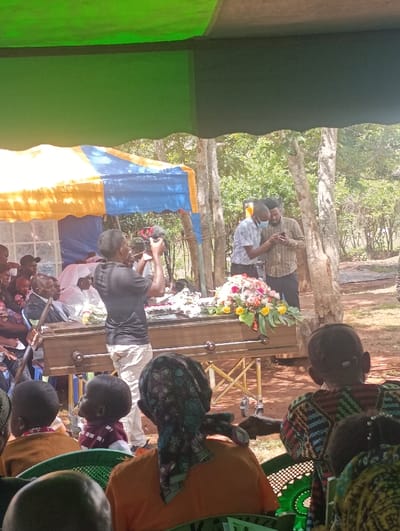 The image portrays an outdoor funeral or memorial service situated in Muthetheni ward, Kenya. The event occurs under a large green canopy, with a smaller yellow and blue striped tent positioned to the left. A brown wooden coffin rests centrally on a gold-colored wheeled stand, heavily adorned with fresh floral arrangements featuring white, pink, yellow, and red flowers on its lid.
In the immediate foreground, several attendees are seated on green plastic chairs with their backs to the viewer. These include an adult female wearing an orange shirt and a multi-patterned headscarf, a young child in a brown top, another child in a maroon and white striped shirt looking towards the coffin, and an adult in a multi-colored patterned garment.
In the midground, a male individual in a black short-sleeved shirt and light-colored pants stands directly behind the coffin, actively operating a black camera pointed towards the coffin and its floral display. To the left of the coffin, under the yellow and blue tent, several individuals are seated, one being an elderly man in a dark suit jacket. To the right of the coffin, two adult males are standing; one wears a light-colored collared shirt, dark pants, and a light blue medical mask, looking at a smartphone held by the other man, who is dressed in a patterned brown long-sleeved shirt.
The environment features reddish-brown earth and a dense background of trees and foliage, indicating a natural, outdoor setting under bright daylight.