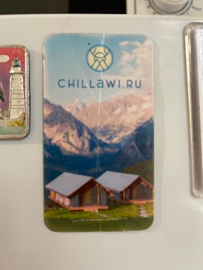 A magnet featuring a scenic mountain landscape and two modern cabins is adhered to a white surface, likely a refrigerator. The magnet has a light blue sky with white clouds, and pastel-colored mountains in the background. Two cabins with dark wood siding and light gray roofs are situated on a grassy hill. A logo with a yellow circular emblem and blue text that reads "CHILLAWI.RU" is positioned in the upper center of the magnet. The magnet shows signs of wear, with visible cracks on its surface. Another magnet, partially visible on the left, depicts a white lighthouse. The overall context suggests the magnet is a souvenir or promotional item. The location in Dolakovo, Russia, is not directly depicted in the image.