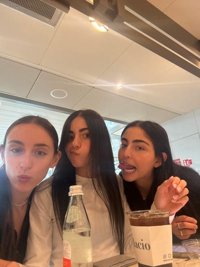 Three young adult females are depicted in a close-up, selfie-style photograph, seated indoors at what appears to be a cafe or restaurant.

From left to right:
The first female, on the left, has dark hair pulled back and wears a dark top with a thin white necklace. She makes a pouting facial expression.
The second female, in the center, has long, straight dark hair and wears a light-colored, long-sleeved top. She also has a pouting expression with slightly wide eyes. In front of her is a clear glass bottle with a silver screw cap, partially filled with water, and a partial red label.
The third female, on the right, has long dark hair and wears a dark top. She is making a playful face, sticking her tongue out. Her right hand, with light pink fingernail polish and a ring on her ring finger, is visible near a clear plastic cup. The cup has a white paper sleeve labeled "Bacio" with an Instagram logo and "MADE IN THE US" text. It contains a dark liquid and ice. A small, transparent glass dish is also visible.

The background shows a modern indoor setting with a light-colored ceiling featuring recessed circular and square lighting fixtures, one with a grate. Dark structural beams are visible in the upper right. Behind the individuals, a large window or glass partition reveals reflections of modern building structures, suggesting an urban or commercial environment. A red sign with white lettering is visible on the far right wall.

The specific location within Berry Islands, Bahamas, is not discernible from the visual evidence present in the image.