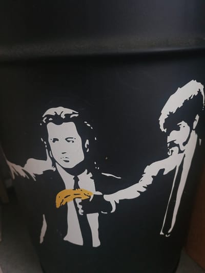 The image shows a stencil art of two men in suits, a famous scene from Quentin Tarantino's film Pulp Fiction. The man on the left is holding a yellow banana, and the man on the right is pointing a weapon towards the viewer. The artwork is on a black surface, likely a trash can or bin, given the curvature of the surface. The artwork is a stencil, suggesting it's not a photograph or a painting. The scene depicts an iconic moment of the film, characterized by the characters' stylized clothing and the presence of the banana as a comedic prop in a tense situation. There is no visible indication of the specific location in the city of Nakhabino, Russia.