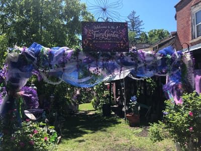 The image shows a whimsical entrance to a fairy garden, adorned with flowing purple and blue fabrics, artificial greenery, and decorative flowers. A sign above the archway reads "Welcome to the Fairy Garden," featuring illustrations of fairies and mushroom houses. Beyond the archway, a grassy path leads towards a rustic shed or building, also decorated with fairy garden elements. The surrounding environment includes lush green trees and a bright blue sky, suggesting a sunny day. The scene is located in the city of Severn, Canada, likely a residential or garden setting designed for imaginative play or decoration. There are no people visible in the image.