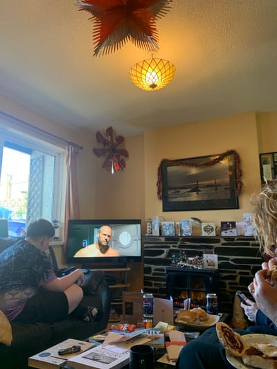 The scene depicts an interior view of a living room in Tideford, United Kingdom. Two individuals are present. On the left, a person wearing a patterned short-sleeved top and dark shorts sits on a dark sofa, facing a television. The television displays an image of a bald man, possibly an actor, against a grey metallic background with a circular window-like feature. Below the television, a dark stand holds various items including a book. On the right, partially out of frame, another individual with curly hair is seated, holding food with both hands while simultaneously gripping a dark smartphone.
The room features pale yellow walls and a white textured ceiling. Two decorative, star-shaped ceiling lights, one predominantly red and silver, and another translucent yellow fixture, illuminate the space. A third smaller, red and silver star-like decoration hangs on the wall near the television. Above a stone-clad fireplace on the right, a large framed picture depicting a coastal scene at dusk or dawn is draped with red tinsel. The mantelpiece holds multiple Christmas cards and small decorative items. An electric fireplace insert glows beneath the mantel.
In the foreground, a dark coffee table is laden with multiple items, including books (one titled "HISTORY"), magazines, assorted papers, snack bags (one labeled "Salty"), two blue Pepsi cans, and a black mug. Plates of food, specifically sandwiches or rolls, some with visible red filling, are also on the table and held by the person on the right. A large window with light-colored curtains on the left provides natural light, revealing an exterior wall or fence structure.