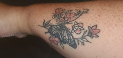 The image shows a tattoo on a person's forearm in Sasbach, Germany. The tattoo depicts a butterfly with intricate wing patterns in black ink. Surrounding the butterfly are several small, stylized flowers in shades of pink and red, with green leaves and branches. One of the flowers, located below and to the left of the butterfly, appears to have a geometric pattern within its petals. The skin tone is light with visible freckles. The background is dark and out of focus, suggesting the image was taken indoors with artificial lighting. The focus is entirely on the tattoo, showcasing its details and colors against the skin. There is no visible text in the image. The lighting creates shadows that highlight the texture of the skin and the tattoo itself.
