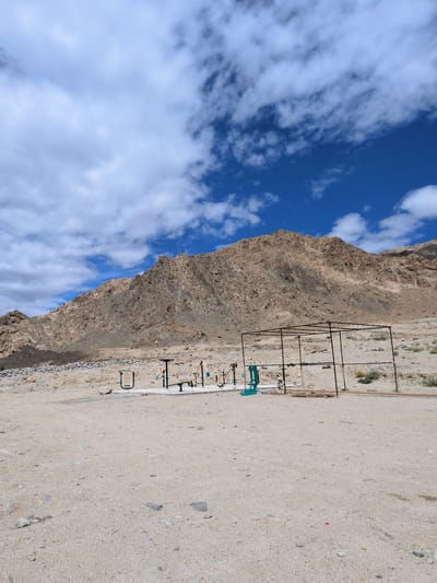 This is a full-shot, eye-level, outdoor photograph taken in Phyang, India. The scene depicts a barren landscape with a large, rocky mountain dominating the background. In the mid-ground, there is a small outdoor exercise area with several pieces of gym equipment, including a pull-up bar, a bench press, and what appear to be leg press machines. There is also a rusted metal frame structure in the mid-ground, possibly a shelter or a partially constructed building. The ground is covered in sand and small stones. The sky is a vibrant blue with scattered white clouds. No people are visible in the image.