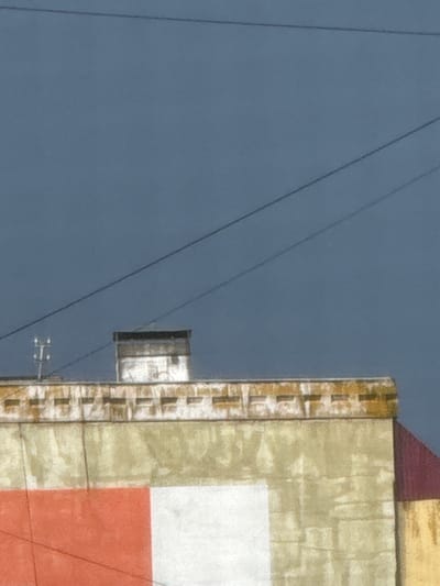 The image shows a close-up of the upper facade of a building against a dark blue sky with thin, dark power lines stretched across it. The building's exterior has a textured appearance, with what looks like brickwork along the top edge, painted in muted yellow and white. Below this, the wall is primarily a pale yellow, with a large section of red on the lower left, and a white section next to it. A red roofline is visible on the right side of the frame. A white chimney with a dark top stands on the roof. On the left side, a small, metallic structure with a "T" shape is visible above the building's facade, likely a utility pole. The scene appears to be in an urban or semi-urban environment in Морозовка, Russia, given the presence of utility infrastructure and building architecture. There are no people visible in the image.