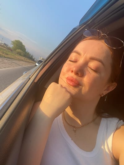 A woman is resting her head on her hand inside a car. She is wearing sunglasses on her head, a white shirt, and a gold necklace with a cross pendant. The car appears to be in motion on a road outside the city of Совхозный, Russia, as evidenced by the passing landscape visible through the car window. The road is bordered by fields and some buildings.