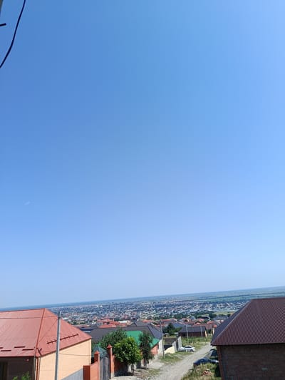 The image is a high-angle, panoramic view of a residential area with houses and a distant cityscape under a clear blue sky. In the foreground, there are several houses with red and brown roofs. One house on the left is brick-colored with a red corrugated metal roof. To its right, there is a light orange brick house with a gray concrete base and a prominent utility pole. Further to the right, another house has a dark brown, corrugated metal roof. A dirt road is visible in the middle ground, with two cars parked on it. Beyond the immediate houses, the image shows a dense expanse of buildings with various colored roofs, extending towards the horizon where it meets a hazy landscape of fields and possibly a body of water. The scene appears to be a suburban or semi-urban environment, possibly in the city of Novyy Benoy, Russia, given the architectural style and landscape. There are no people visible in the image.