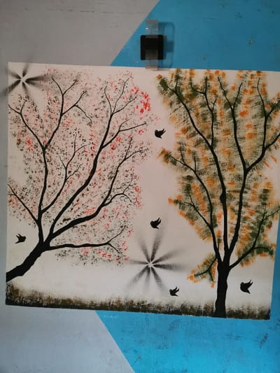 The image shows a wall painting of two trees with black silhouettes against a white background. The tree on the left has pink, red, and brown leaves, while the tree on the right has yellow, orange, and brown leaves. There are several black birds painted on the white space between the trees, as well as a spray-painted starburst design. The painting is located in the town of Gerin Gada, Nigeria, which has the coordinates latitude: 9.93, longitude: 8.86. The painting is mounted on a wall, and it is likely located in a public area. The painting is made of paint, and it appears to have been painted recently.  The colors are vibrant and the details are precise, suggesting that the artist was skilled. The painting may be a symbolic representation of nature and beauty.