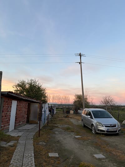 The image depicts a rural scene in Liahovo, Bulgaria, taken during a sunset. A silver Opel Zafira with license plate PA 6774 KX is parked on a dirt and gravel area. Overhead power lines are strung from a concrete utility pole. Adjacent to a paved pathway is a low, brick structure with a tiled roof. Clothes are hanging on a clothesline. The sky shows colors ranging from light blue to pink and orange. Vegetation consists of trees and dry grass.