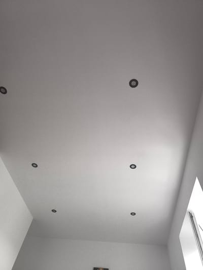 The image depicts a white ceiling with several recessed light fixtures. There are six such fixtures visible in the frame, evenly spaced across the surface of the ceiling. The lighting appears to be dim, casting subtle shadows. In the lower right corner, a portion of a white wall and a window frame are visible, suggesting an indoor setting. The image is a high-angle shot, looking upwards. There is no indication of the location being in Katowice, Poland.