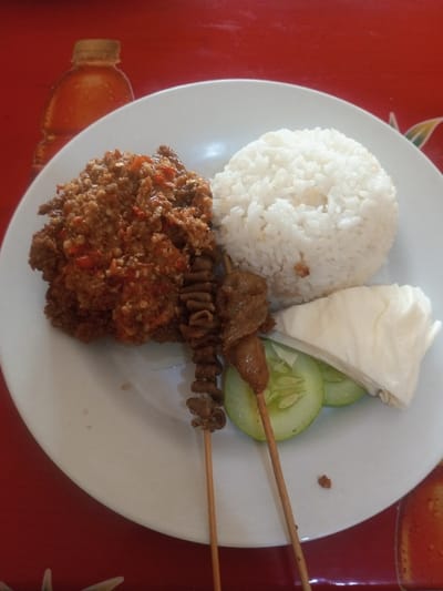 A white plate holds a serving of rice molded into a dome shape. To the left, a pile of what appears to be ground meat is heavily seasoned with red chilies. Two skewers of meat rest on the plate, alongside cucumber slices and a piece of cabbage. The red surface underneath features a faded pattern including a juice bottle graphic.