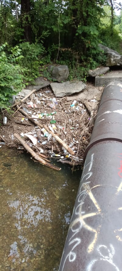 The image shows a waterway clogged with trash and debris in what appears to be a natural setting. A large metal pipe, covered in graffiti, runs along the right edge of the frame, partially submerged in the water. The water is clear enough to see the rocky bed below. A large amount of garbage, including plastic bottles, cans, and general waste, is tangled with branches and other natural debris, forming a significant blockage in the stream. The banks of the stream are lined with lush greenery, including trees and shrubs. The setting is a natural area near an urban one, with the large metal pipe being a human-made element. The city is Tahlequah, United States of America.