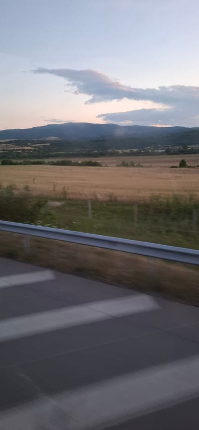 The image depicts a rural landscape at dusk, possibly near Mursalevo, Bulgaria. The foreground features a blurred road surface with white stripes, suggesting a perspective from a moving vehicle. A silver metal guardrail separates the road from a field of dry, golden grass that extends into the mid-ground. Beyond the field, a dark, tree-covered hillside rises, leading to a range of hazy, blue mountains in the distance. The sky above is a pale, dusky blue, with scattered, wispy clouds tinged with pink and gray. The overall impression is one of a serene, open landscape captured during the transition from day to night.
