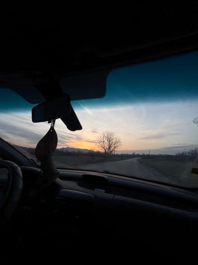 The image presents a forward-facing view from inside a vehicle on a paved road through a rural landscape in Gorna Rositsa, Bulgaria, under twilight conditions consistent with sunset or sunrise. The vehicle's interior, including the roof lining, dashboard, and a portion of the steering wheel on the left, is largely in shadow. A rearview mirror hangs centrally, with a dark reddish-brown air freshener, possibly leaf-shaped, suspended from it. A blue-tinted sun visor strip is visible across the top of the windshield, and a white suction cup mount is affixed to the upper right corner of the windshield.
Through the windshield, a two-lane road curves gently to the right, receding into the distance. The horizon on the left exhibits an orange and yellow hue, indicating the sun's position, fading into light blue and greyish-white clouds higher up and to the right. A faint contrail is visible in the upper sky. A prominent, bare deciduous tree stands silhouetted against the illuminated horizon, to the left of the road. Distant mountains are visible along the horizon line. The road is flanked by fallow fields, and utility poles with wires are discernable along the right side of the road further in the distance.