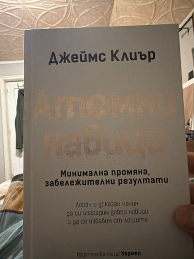 The scene depicts a paperback book held by a human hand, positioned prominently in the foreground. The book has a gray cover with text in Bulgarian. The author's name, "Джеймс Клиър" (James Clear), is printed in black at the top. The title, "АТОМНИ НАВИЦИ" (Atomic Habits), is rendered in a large, stippled, light orange-gold font below the author. Further text in black includes the subtitle "Минимална промяна, забележителни резултати" (Minimal change, remarkable results) and a descriptive phrase "Лесен и доказан начин да си изградим добри навици и да се избавим от лошите" (An easy and proven way to build good habits and get rid of bad ones). The publisher, "Издателска къща Хермес" (Hermes Publishing House), is indicated at the bottom. A light-toned human hand is partially visible on the right side, gripping the book. The background includes an ornate, light-colored patterned ceiling or wall in the upper left. To the left, a light-colored door frame or window frame is partially visible, with a dark garment hanging on a hook behind it. The lower left shows blurred textiles, possibly bedding, in warm tones. The lighting suggests an indoor setting. This scene is located in Drachevo, Bulgaria.