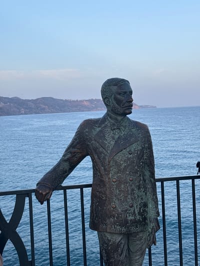 A bronze statue of a man in a suit and bow tie stands on a railing overlooking the ocean. The man is leaning his left arm on the railing. The statue is green with age. In the background, there are mountains and the sea. The sky is a light blue with some clouds. The railing is black and ornate. The statue is in the town of Nerja, Spain.