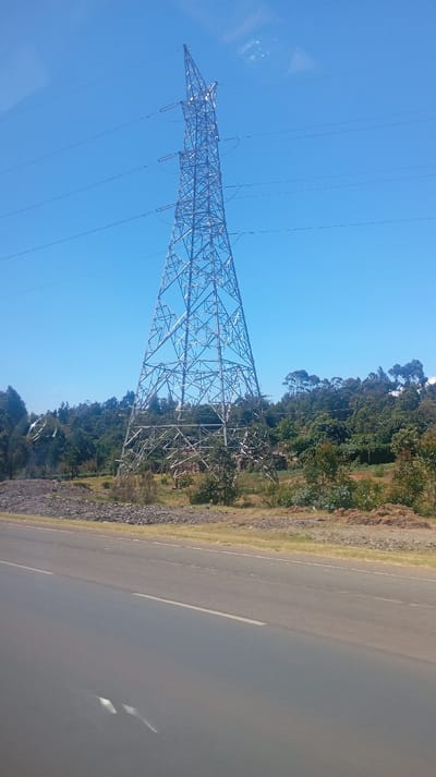 The image displays an outdoor scene featuring a large electricity transmission pylon positioned prominently next to a road, set against a backdrop of lush vegetation and a clear blue sky.
A tall, metallic lattice-work electricity pylon stands in the mid-ground on the left side of the frame. Its structure is made of numerous interconnected steel beams that form a wide, stable base, tapering significantly towards its pointed top. Multiple high-voltage electrical transmission lines extend horizontally from the pylon, secured by white disc-shaped insulators.
The sky is a clear and vibrant blue, indicating bright, sunny weather. There are no distinct clouds, though some faint, diffused white smudges or reflections are visible in the upper right quadrant, possibly reflections from a car window.
Behind the pylon and extending across the middle and right side of the image, a dense expanse of green trees and bushes forms a natural background. The vegetation appears healthy and varied, suggesting a forested area, a hillside, or well-vegetated terrain. Patches of lighter ground or smaller clearings are discernible among the trees.
The ground around the base of the pylon and extending towards the road is uneven, covered with dry grass, patches of brown earth, and scattered rocks or debris. Some smaller bushes and sparse vegetation grow in this area.
In the foreground, a dark grey asphalt road stretches horizontally across the bottom portion of the image. It appears to be a paved highway or main thoroughfare with distinct lane markings. A continuous white line marks the edge of the road on the far right, while a dashed white line separates lanes on the left side of the visible road. The road surface shows some reflective qualities in the lower left, and blurred white markings, along with indistinct reflections, suggest the photograph was taken from inside a moving vehicle, looking out a window.
The scene is brightly lit by natural sunlight, indicating it was taken during the day. There is no visible text in the image. The overall impression is of a journey through a rural or semi-rural landscape.