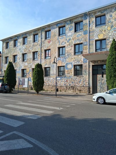 The image displays a vibrantly decorated building with a mural covering its facade. The artwork features a playful and abstract style with a variety of shapes, symbols, and cartoonish figures in yellow, blue, and beige tones. Several windows are visible on the building's multiple stories. In front of the building, a pedestrian crossing with white stripes is marked on the asphalt road. A white car is parked on the right side of the road, partially visible. Two dark green, rounded bushes are planted in front of the building, flanking the pedestrian crossing. A lamppost stands near the building, and a trash can is visible next to it. Two posters are displayed on stands in front of the building, one featuring a black and white portrait and the other with text. The sky is a clear blue, suggesting fair weather. The scene appears to be a street in a town or city, likely during the daytime. The overall impression is one of urban art and public space.