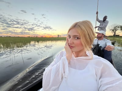 A woman poses in the foreground of a selfie photo, while two men are seated behind her in what appears to be a boat traveling on a river or waterway. The location is likely within the city of Lobilos, Botswana, as per the prompt instructions.

The woman is fair-skinned with blond hair styled in an updo. She wears a white blouse and has her left hand raised to her cheek in a posing gesture. She wears a ring.

The man behind her, closest to the camera, has dark hair and a beard, wears a white baseball cap, and a light blue button-down shirt. Binoculars hang from his neck.

The third man stands at the rear of the boat, using a pole to propel it through the water. He is Black, tall, and dressed in a grey button-down shirt.

The setting is an open river or waterway with tall grasses visible along the banks and a sunset or sunrise sky with scattered clouds in the background. The water reflects the sky and vegetation. The boat is dark in color.