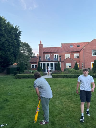 This image captures a residential outdoor scene in Bubwith, United Kingdom, during late afternoon or early evening. The foreground features a well-maintained green lawn.
Two male subjects occupy the foreground. The first, positioned left of center and viewed from behind, is a light-skinned individual with brown, wavy hair. He wears a light-colored checkered short-sleeved shirt, olive-green trousers, and burgundy New Balance sneakers. He holds a yellow cricket bat with blue, red, and white decals in a ready stance, pointing towards the background. The second male subject, positioned right of center, is light-skinned with dark, upward-styled hair. He wears a white t-shirt, dark shorts, white socks, and black and white sneakers, and is depicted walking towards the right side of the frame.
In the mid-ground, a manicured garden area with low hedges and conical topiary shrubs separates the lawn from a large, multi-story brick house. The house features a red tiled roof, multiple white-framed windows, a second-story balcony with a dark railing, and extensive ground-floor glass doors. Through these glass doors, at least two additional individuals are visible; one is discernibly holding a cricket bat, suggesting a casual game is underway. The house exhibits a prominent brick chimney on its left side and a skylight on the right section of the roof.
Large, dark green evergreen trees flank the left side of the scene, extending upwards into a clear, light blue sky that displays a faint pink-orange tint near the horizon. A second cricket bat is partially visible on the grass in the far-left foreground. The overall scene depicts recreational activity within a private, well-maintained residential property.