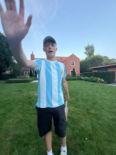 This image captures a young male in a full-body, front-facing perspective, standing on a well-maintained green lawn. He is wearing a green baseball cap, a light blue and white vertically striped t-shirt featuring a crest on the left chest (resembling an Argentina national football team jersey), black cargo shorts, white socks, and white athletic shoes. His left hand is extended directly towards the camera with the palm open, while his right hand holds a light-colored wooden cricket bat positioned alongside his right leg. The subject's mouth is slightly open. In the background, a two-story residential house constructed of red brick with white-framed windows and a red roof is visible. To the right of the house, a single-story modern structure with horizontal wooden paneling and dark window panes stands. The property is characterized by manicured hedges, assorted trees, and landscaped shrubbery. The sky above is clear blue with minimal cloud cover. The scene is located in Bubwith, United Kingdom.