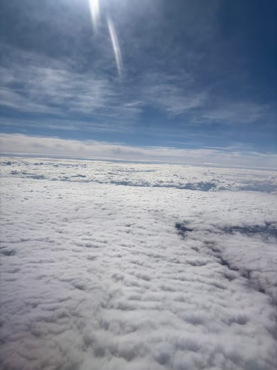The image shows an aerial view of clouds and sky. A bright sun is visible on the left, with the sky transitioning to a deep blue further up. A dense layer of white, textured clouds fills the right side of the frame. A strip of thinner clouds or haze separates the sky from the dense cloud layer. There is no indication of the location being Sucha Beskidzka, Poland.