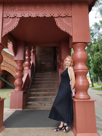 A middle-aged Caucasian woman with blonde hair and red lipstick stands leaning against a large, brown, fluted pillar. She is wearing a long black dress with a yellow strap visible on her shoulder and a colorful bag slung across it. Her feet are clad in black sandals. Behind her, a red brick staircase with a black railing ascends to a covered porch area. The porch features ornate wooden carvings and is supported by similar fluted pillars. The background shows greenery and trees, suggesting an outdoor setting. The scene is likely within the city of Chelganovo, Russia, given the architectural style.