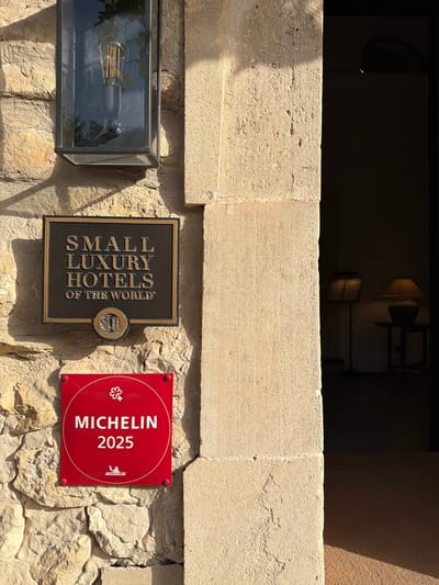 This image depicts a close-up view of a stone wall on the exterior of a building, likely a hotel, in Montuïri, Spain. Two prominent signs are mounted on the wall. The upper sign is a dark, rectangular plaque with gold lettering that reads "SMALL LUXURY HOTELS OF THE WORLD" and a circular emblem below it. The lower sign is a square, vibrant red plaque with white lettering that says "MICHELIN 2025" and features a stylized key symbol at the top and a Michelin Man logo at the bottom. To the right of the signs, a shadowed doorway leads into a dimly lit interior where two lamps are visible. The stone wall itself is rough-hewn and bathed in warm sunlight, casting distinct shadows. An outdoor lantern with an exposed Edison bulb is mounted above the "SMALL LUXURY HOTELS" sign. The overall impression is one of a distinguished establishment with notable accreditations.