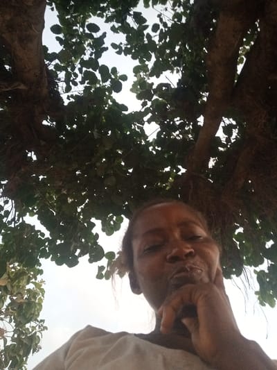 A low-angle shot features a woman gazing downward, her hand resting on her chin. The light is subdued, emphasizing her features. Above her, a dense canopy of tree branches and leaves fills most of the frame, partially obscuring the sky. Another patch of leafy vegetation is visible on the left side. The overall composition creates a sense of being surrounded by nature.