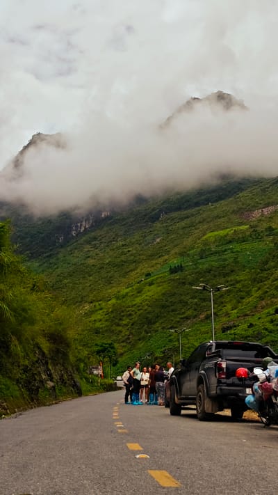 The image depicts a scene on a winding road in the Phố Bảng Commune, Vietnam, with a steep, green, mountainous backdrop partially obscured by low-hanging clouds. A group of approximately seven people stands on the road near a black pickup truck. Some members of the group are wearing blue protective coverings over their shoes. A motorcycle with several helmets is parked to the right of the truck. The road features a dotted yellow center line. The weather appears overcast and damp. Streetlights are visible in the background along the road. Vegetation flanks the road.