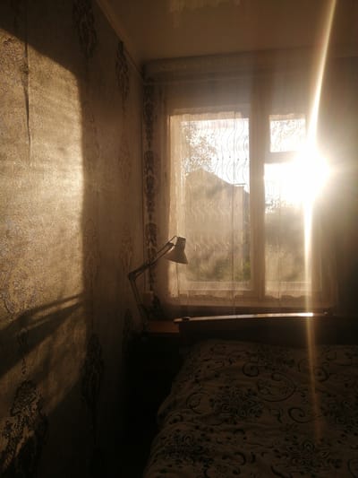 The image depicts an interior scene, likely a bedroom, in Dubrovka, Russia. The primary focus is the interplay of light and shadow within the room. A strong beam of sunlight streams in from a window, illuminating the right side of the frame and casting dramatic shadows on the patterned wallpaper to the left. The wallpaper features an ornate, damask-like design in muted tones of gold and brown.
Through the sheer, patterned curtains of the window, an outdoor scene is vaguely visible, suggesting houses and foliage, indicating it is daytime. A desk lamp with a flexible arm is positioned on a shelf or table beneath the window, its metallic surface catching some of the light. In the foreground, the corner of a bed is visible, covered with a blanket or comforter that also has a dark, floral pattern.
The intense sunlight suggests it is either early morning or late afternoon. The mood is somewhat serene, with the warm light creating a cozy and perhaps slightly nostalgic atmosphere. There is no visible text in the image. The scene captures a quiet moment, perhaps just waking up or winding down for the day, with the sunlight being the most dynamic element.
