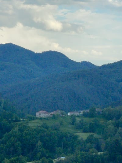 The image depicts a mountainous landscape under a cloudy sky. A mountain range covered in dense, dark green vegetation dominates the right side of the image.  A building with light-colored walls and dark roof can be seen in the middle ground of the right side. The location of this image is in Zhaltusha, Bulgaria.