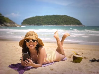 A young Asian woman with long dark hair, wearing a straw hat, white cat-eye sunglasses, and a yellow and white striped one-piece swimsuit, is lying on a beach. She is propped up on her elbows, holding a black smartphone in her hands, and appears to be listening to something through white earbuds. She is smiling and looking towards the camera. Beside her, on the sand, is a whole coconut with a yellow straw, and a pair of black-rimmed glasses. The beach has light-colored sand, and the ocean with waves is in the background, with a small, tree-covered island in the distance under a clear blue sky. The scene suggests a tropical vacation setting. There is no indication that this image is in Bevern, Germany.