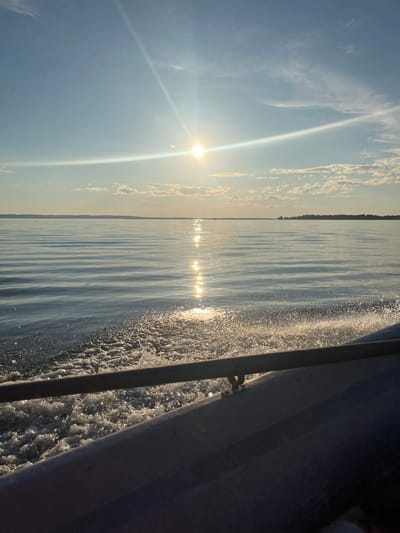 The image captures a serene scene from a boat moving across a body of water, likely in Siukterka, Russia. The sun, positioned high in the sky and casting a brilliant glare, suggests it is midday. Wispy clouds streak across a clear blue sky. The water's surface is calm, with gentle ripples creating a textured pattern. Sunlight reflects off the water, forming shimmering lines that stretch towards the horizon. The distant shoreline is visible as a dark, silhouetted line, hinting at land or a forest. The right side of the frame is dominated by the side of the boat, with frothy white water churning in its wake, indicating movement. The overall mood is peaceful and tranquil, evoking a sense of calm and the beauty of nature on a sunny day. There is no visible text in the image.
